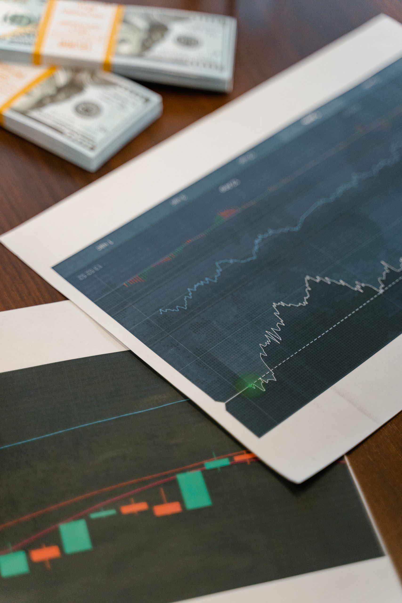 Papers with financial graphs beside stacks of US dollar bills representing market analytics.