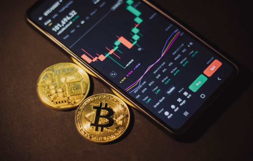 Smartphone displaying trading app with Bitcoin coins on a dark surface.