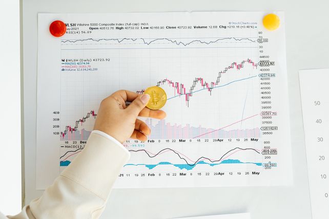 A hand holding a Bitcoin coin in front of a stock market chart, symbolizing analysis and finance.