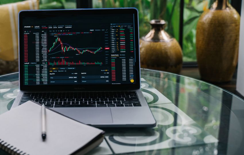 Laptop displaying cryptocurrency stocks and graphs on a glass table with a notepad.