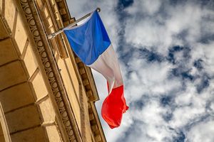 The French flag waves against a cloudy sky, mounted on a Parisian building.