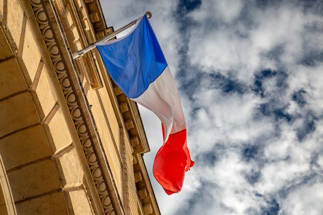 The French flag waves against a cloudy sky, mounted on a Parisian building.