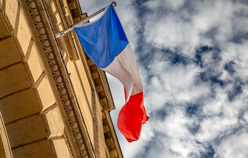 The French flag waves against a cloudy sky, mounted on a Parisian building.