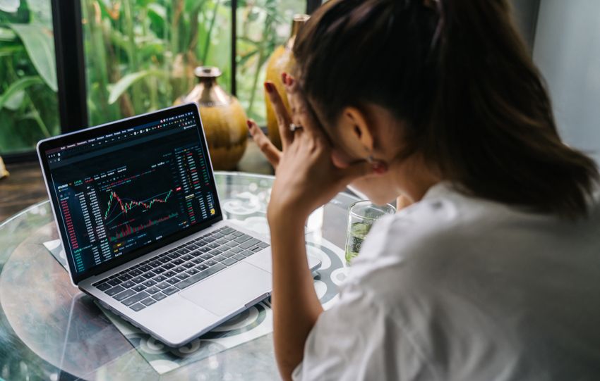 Woman analyzing cryptocurrency market charts on a laptop, looking stressed in a cozy indoor setting.