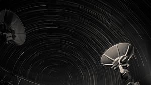 Long exposure of star trails creating circular patterns over two satellite dishes against a night sky.