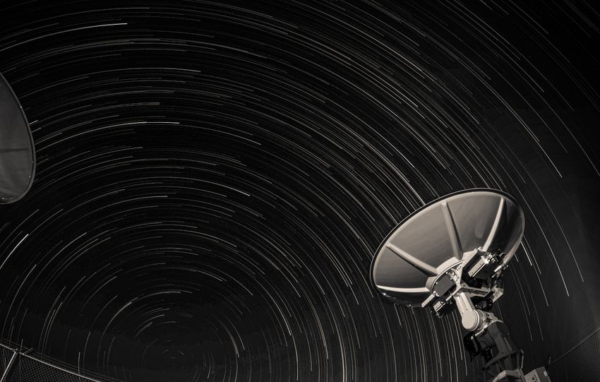 Long exposure of star trails creating circular patterns over two satellite dishes against a night sky.