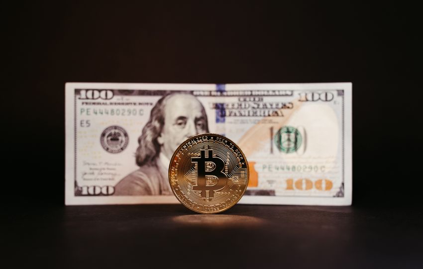 Bitcoin coin standing in front of a 100 dollar bill, symbolizing cryptocurrency and traditional currency themes.